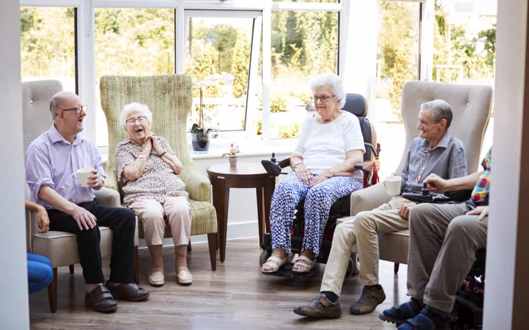 Male And Female Residents Sitting In Chairs And Talking In Lounge Of Retirement Home