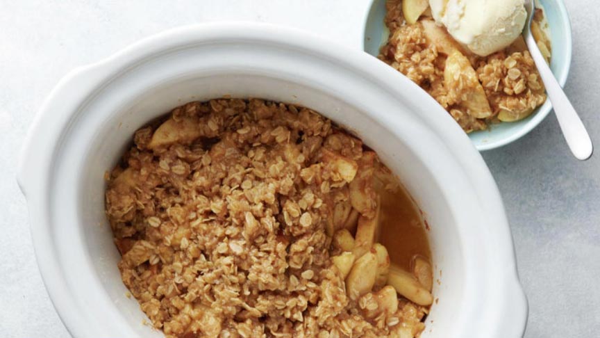 A slow cooker filled with apple crisp, topped with oat crumble. Beside it, a bowl holds a serving of the dessert with a scoop of vanilla ice cream. A spoon rests in the bowl.