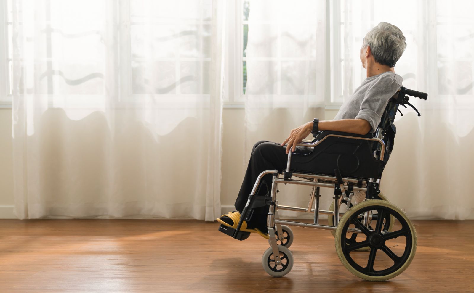 An older man sitting in a wheelchair looking out a window.