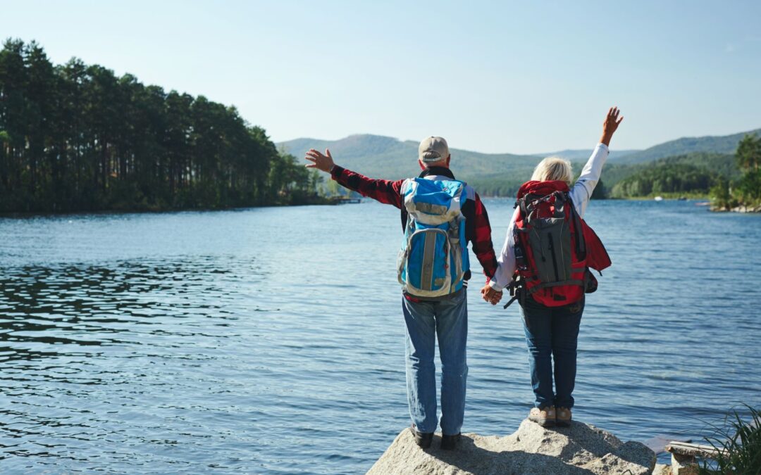 Seniors traveling, with their arms up in the air in front of a large lake.