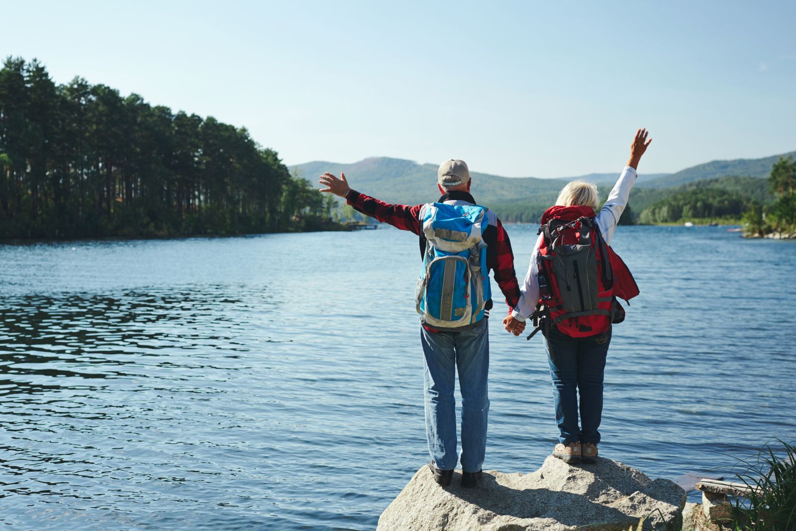 Seniors traveling, with their arms up in the air in front of a large lake.