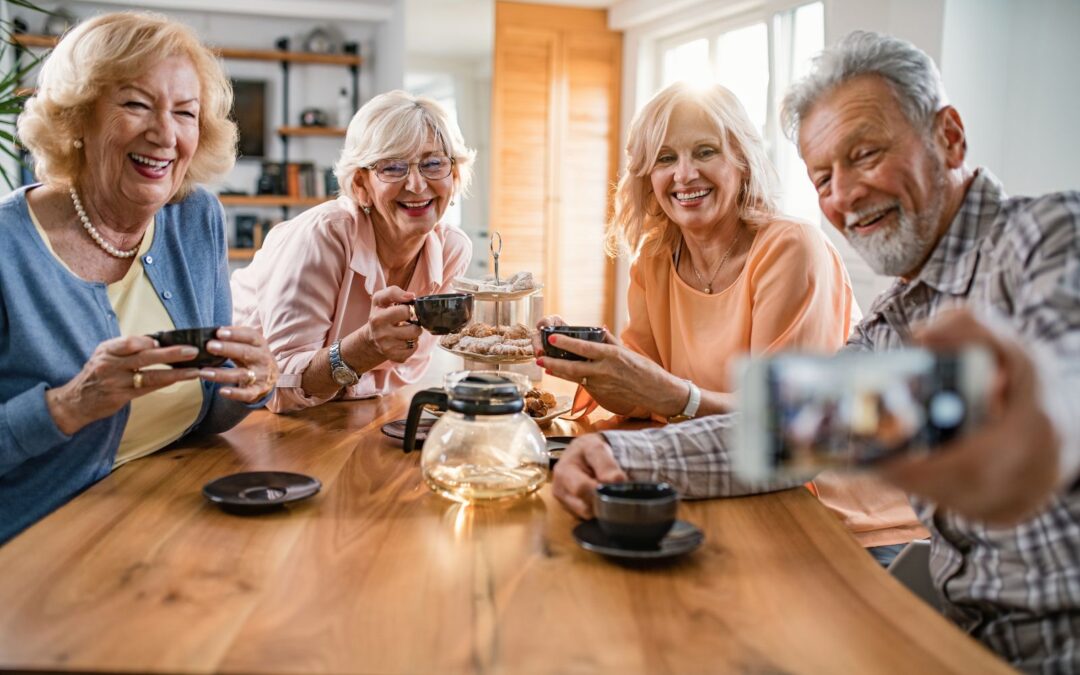 Four senior citizens sitting around a table smiling, taking a selfie on a mobile phone.