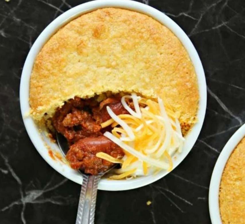 A bowl of chili topped with shredded cheese and covered with a layer of baked cornbread. A spoon is scooping out a portion, revealing the chili beneath. The bowl rests on a dark marble surface.