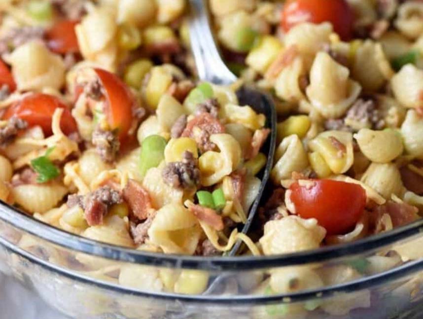 A glass bowl filled with a pasta salad featuring shell pasta, cherry tomatoes, corn, bacon, ground meat, and green onions. A spoon is placed in the salad, showcasing the colorful ingredients.