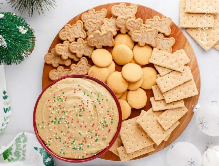 A round wooden board filled with gingerbread cookies, vanilla wafers, and graham crackers surrounding a bowl of creamy dip topped with colorful sprinkles. Holiday decorations like greenery and white ornaments are in the background.