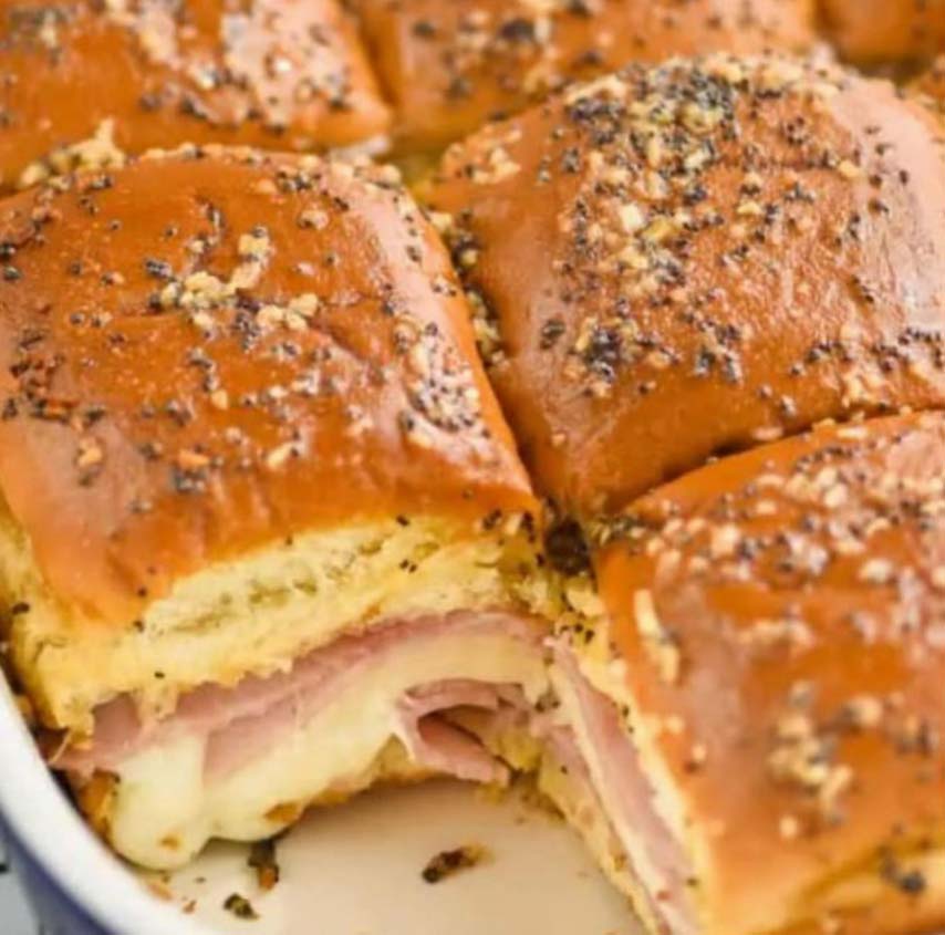 Close-up of baked ham and cheese sliders in a baking dish. The sliders have golden brown tops seasoned with herbs and chopped onions, showcasing layers of melted cheese and sliced ham between soft, fluffy buns.
