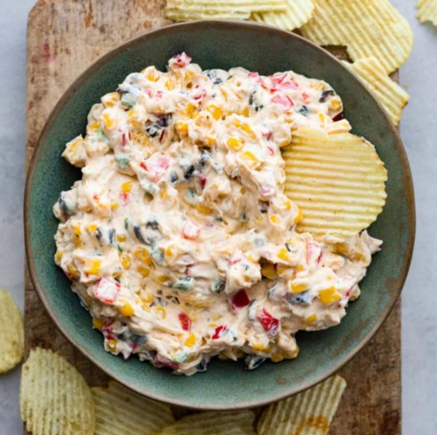A bowl of creamy dip with chopped vegetables, including red bell peppers and corn, garnished with a ridged potato chip. The dip is placed on a wooden board, surrounded by more potato chips.